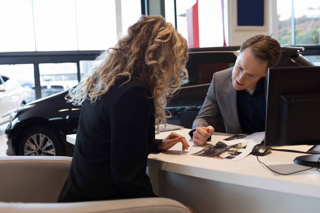  A man and a woman are sitting at a desk in a car dealership. The woman is looking at a brochure, and the man is pointing at something on the computer screen. The image represents the search query 'Selling financed goods that are still unpaid'.